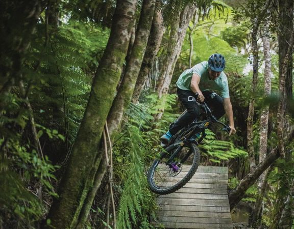 A downhill mountain bike rider going over a wooden drop jump on Dozer trail in Wainuiomata.