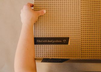 A person's left hand touches the top of a carboard box that has a black "XO" pattern and the words "Filled with local goodness." printed on it.
