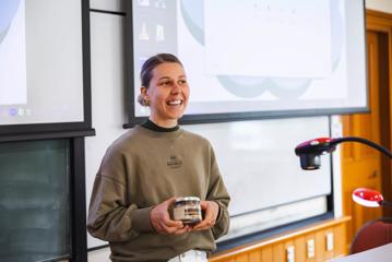 A marketing student at Victoria University stands at the front of a lecture hall holding a glass jar of cacao husk tea from be happy chocolate.