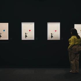 A person wearing a green shirt and leopard print pants stands with their hands in their pockets looking at Banksy's Girl with Balloon print series in a dark space.