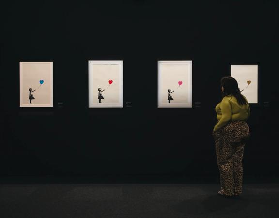 A person wearing a green shirt and leopard print pants stands with their hands in their pockets looking at Banksy's Girl with Balloon print series in a dark space.