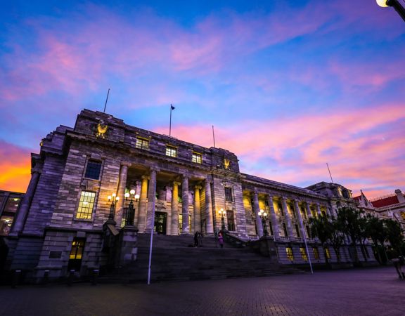 The exterior of the New Zealand Parliament building at 1 Museum Street, Pipitea in Wellington at sunset with pink-coloured clouds in the sky above.