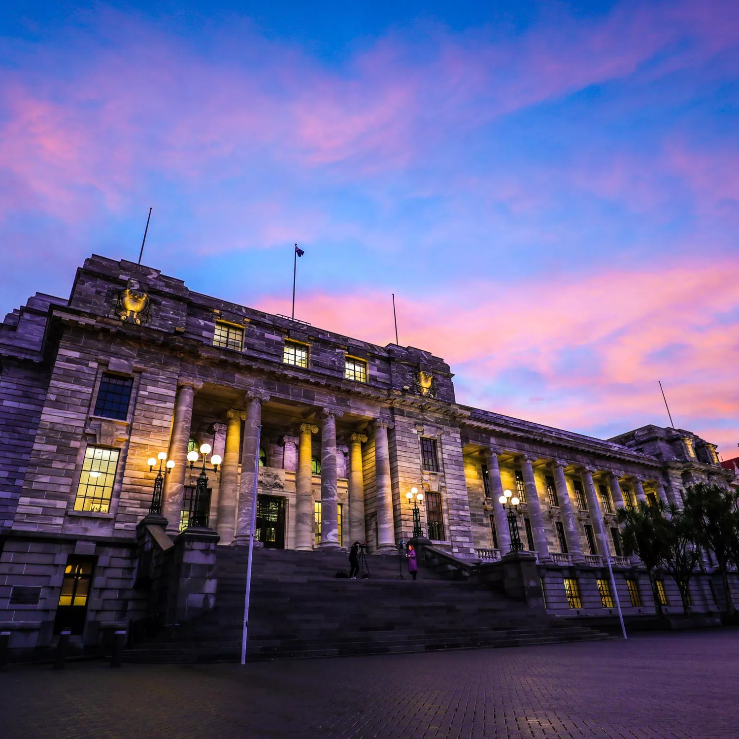 The exterior of the New Zealand Parliament building at 1 Museum Street, Pipitea in Wellington at sunset with pink-coloured clouds in the sky above. 