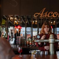 A bartender at Ascot pours a sangio. Large mirrors on the wall behind them reflect outside, and the sign hangs above their head.