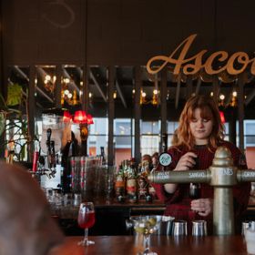 A bartender at Ascot pours a sangio. Large mirrors on the wall behind them reflect outside, and the sign hangs above their head.