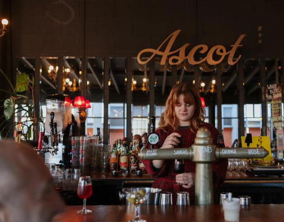 A bartender at Ascot pours a sangio. Large mirrors on the wall behind them reflect outside, and the sign hangs above their head.