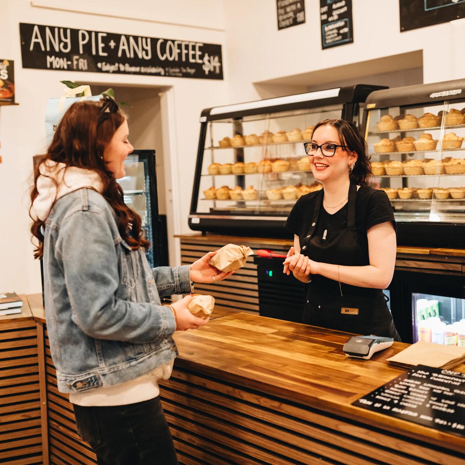 A smiling worker in a black apron sells a couple of pies to a customer.