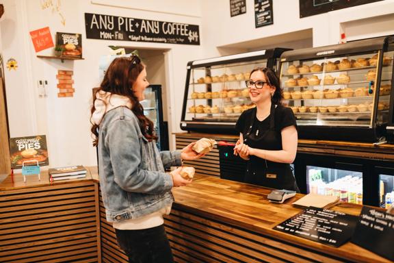 A smiling worker in a black apron sells a couple of pies to a customer.