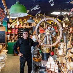 A person standing among a large array of nautical antiques, holding a large ship steering wheel, inside Shipwreck Trading Lower Hutt.