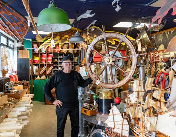 A person standing among a large array of nautical antiques, holding a large ship steering wheel, inside Shipwreck Trading Lower Hutt.