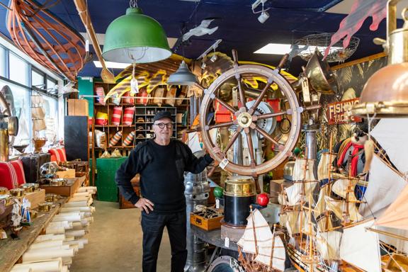 A person standing among a large array of nautical antiques, holding a large ship steering wheel, inside Shipwreck Trading Lower Hutt.