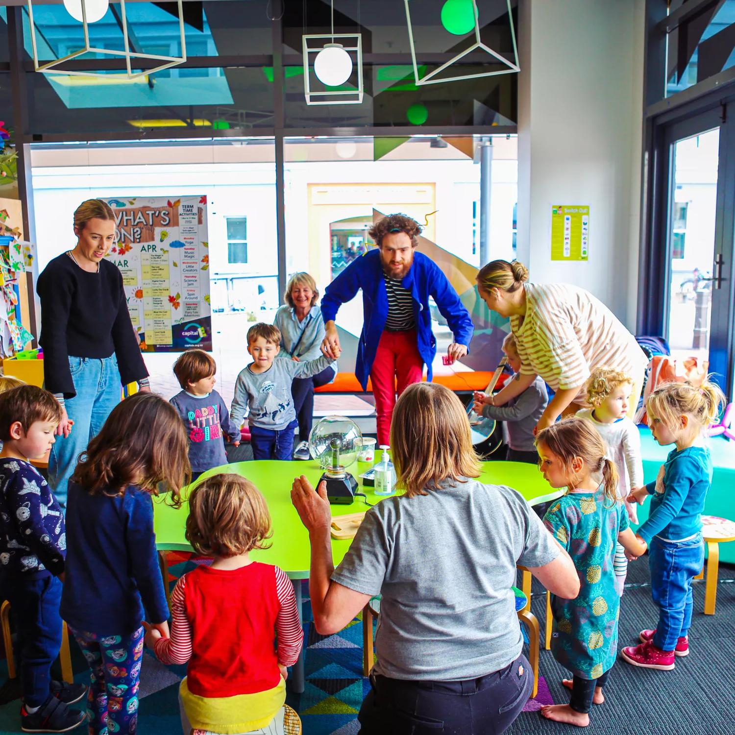 A group of young children with teachers standing around a green table at Capital E, holding hands.