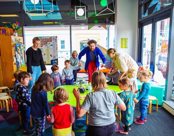 A group of young children with teachers standing around a green table at Capital E, holding hands.