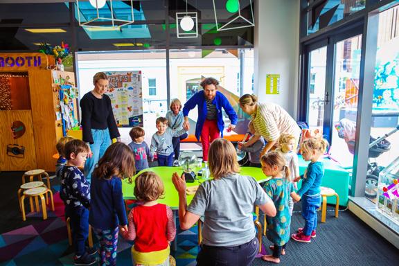 A group of young children with teachers standing around a green table at Capital E, holding hands.