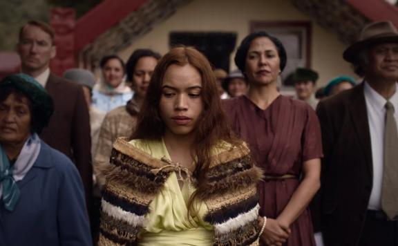 A still from the movie 'Cosuins' where a young person is seen wearing a cloak of honour (Korowai) with their elders standing behind them at a meeting house (Marae).