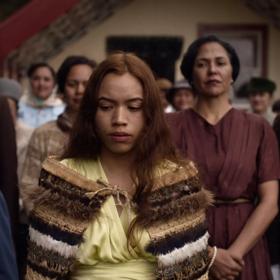 A still from the movie 'Cosuins' where a young person is seen wearing a cloak of honour (Korowai) with their elders standing behind them at a meeting house (Marae).
