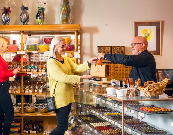 A person wearing a yellow sweater is buying something at Schoc Chocolates, a  chocolatier in Greytown, Wairarapa. The worker is handing them the bag as another shopper is browsing in the background.