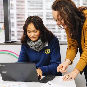 A student uses a laptop computer and a smiling teacher stands by their side.