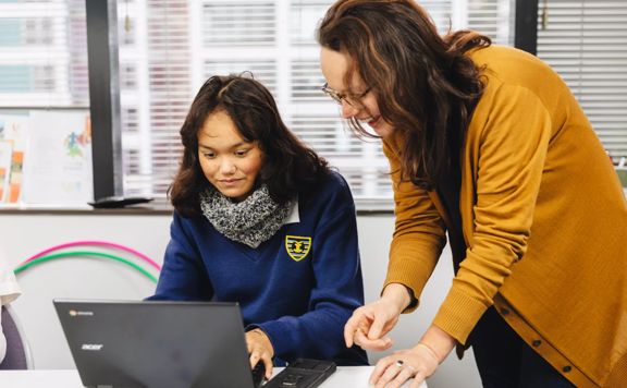 A student uses a laptop computer and a smiling teacher stands by their side.