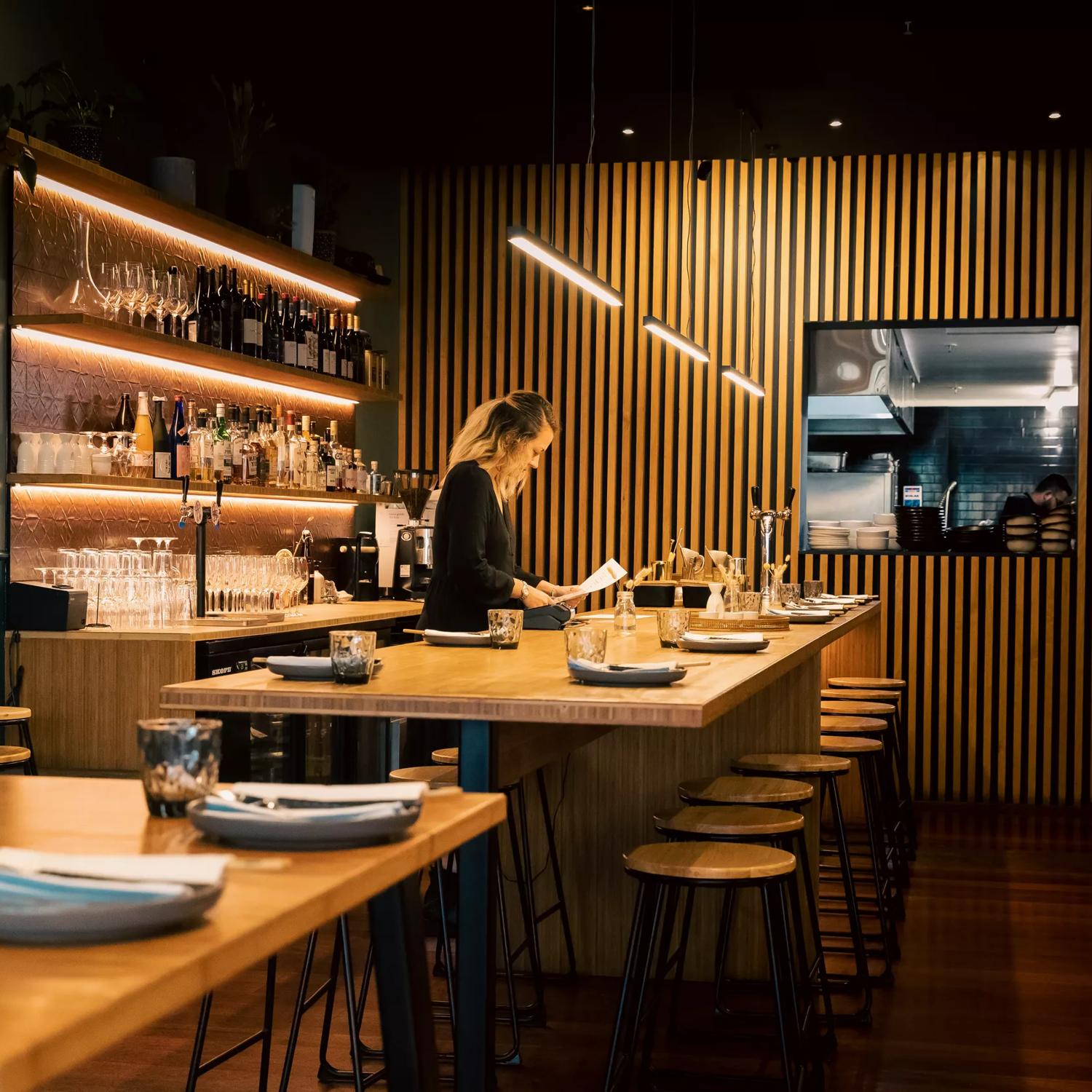 Waiter setting a table at Koji, the room has dim lighting and a large wooden feature wall.