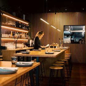 Waiter setting a table at Koji, the room has dim lighting and a large wooden feature wall.