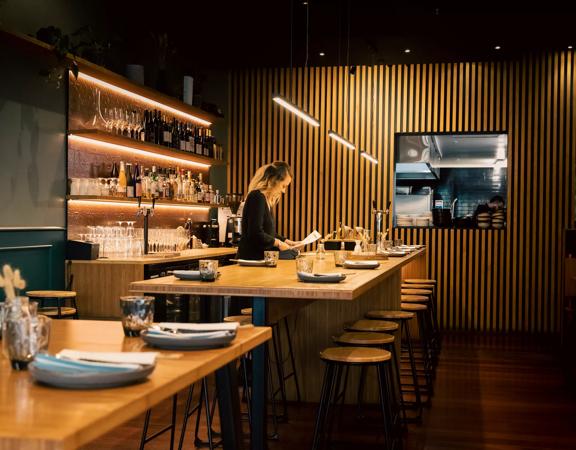 Waiter setting a table at Koji, the room has dim lighting and a large wooden feature wall.