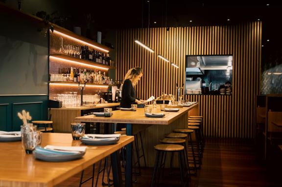 Waiter setting a table at Koji, the room has dim lighting and a large wooden feature wall.