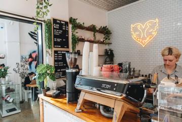 A worker behind the counter at Evil Twins Coffee, a café in Te Aro, Wellington. The bright space has white walls, green plants and a neon sign in the shape of a heart.