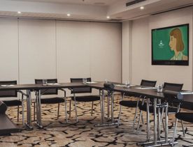 The Bowen Room, a conference room inside the Bolton Hotel in Wellington is setup with the tables arranged in a U shape with six chairs, light grey walls and a mounted TV in the background.