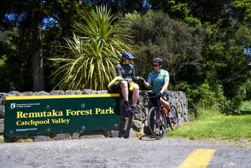 Two cyclists sitting to the left of a Department of Conservation sign for Remutaka Forest Park, Catchpool Valley. Native bush is in the background.