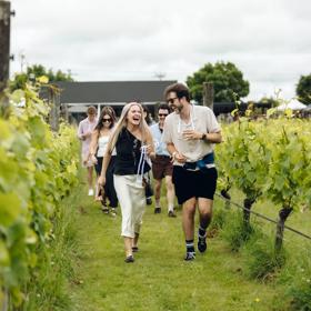 A group of people laughing and drinking while walking through a vineyard.