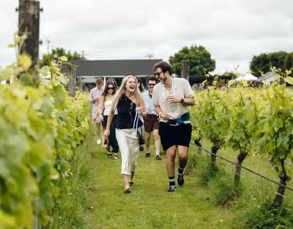 A group of people laughing and drinking while walking through a vineyard.