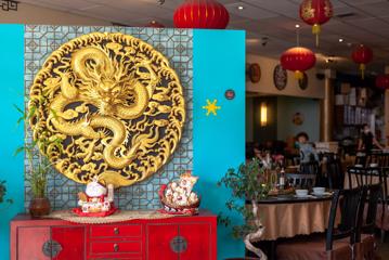 The entrance to a Chinese restaurant. There is a blue wall with a circular basrelief of a golden dragon, a couple of potted plants, and red cabinets with tables and chairs in the background.