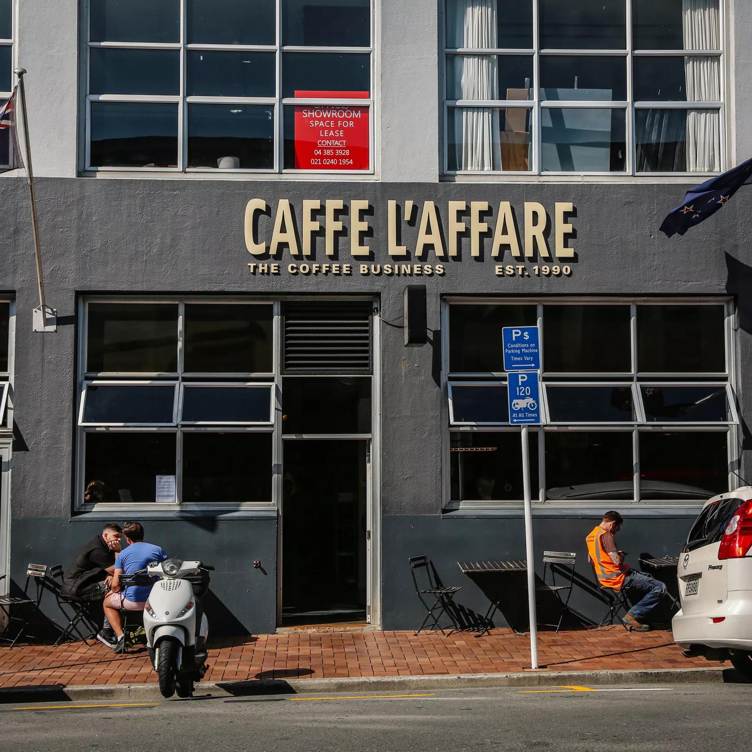 The front facade of Caffe L'Affare located at  27 College Street in Te Aro, Wellington. It's a dark grey cement building with a few people sitting at the small tables outside. 