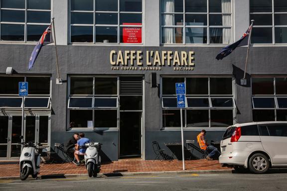 The front facade of Caffe L'Affare located at  27 College Street in Te Aro, Wellington. It's a dark grey cement building with a few people sitting at the small tables outside.