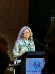 A speaker stands at a podium delivering a talk at a conference, with an IPRES 2025 Wellington logo visible and an audience in the foreground.