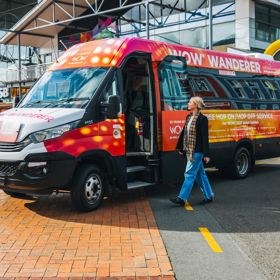 A person walking toward a colourful minibus, covered with World of WearableArt branding and text.
