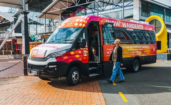 A person walking toward a colourful minibus, covered with World of WearableArt branding and text.