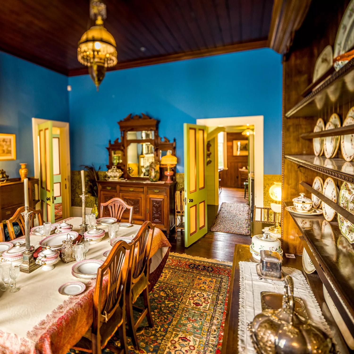 Inside the dining room of the Katherine Mansfield House on Tinakori Road. Blue walls are covered in decor from the 19th century, including a wall of china plates. The table is set with candleabra and pink china.