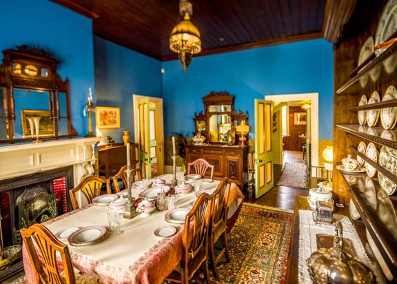 Inside the dining room of the Katherine Mansfield House on Tinakori Road. Blue walls are covered in decor from the 19th century, including a wall of china plates. The table is set with candleabra and pink china.