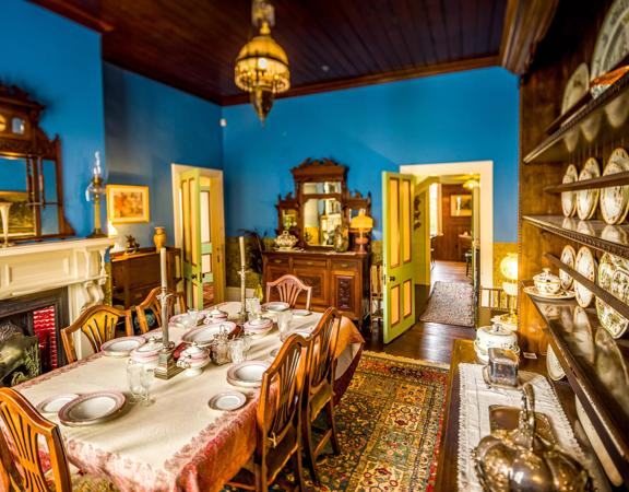 Inside the dining room of the Katherine Mansfield House on Tinakori Road. Blue walls are covered in decor from the 19th century, including a wall of china plates. The table is set with candleabra and pink china.