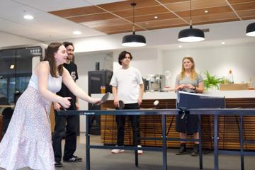 Woman with brown hair, wearing a white shirt and long floral skirt, plays ping pong in a modern office kitchen with three colleagues standing in the background.