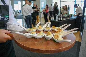 11 large white spoons placed on chopping board with food inside, people blurred in the background talking to eachother.