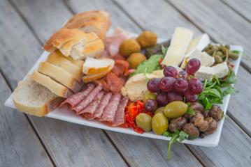 A charcuterie plate with bread, deli meats, hummus, pickled peppers, green olives, grapes, and cheese.