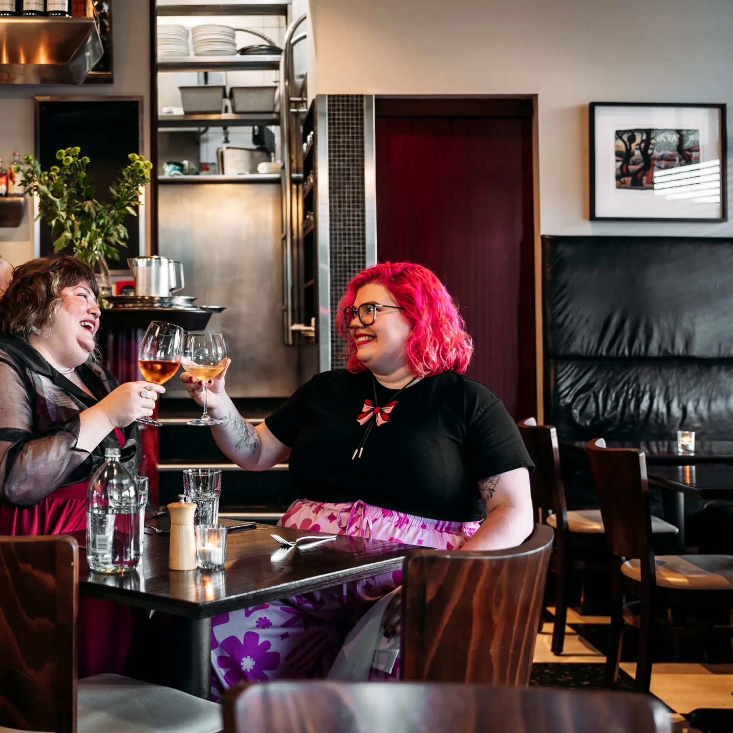 Two people enjoying a meal and clinking their wine glasses in Capitol Restaurant. Waitstaff make drinks behind the bar behind them. The dark wood tables add a moody vibe to the interior.