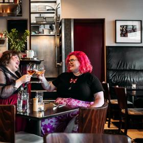 Two people enjoying a meal and clinking their wine glasses in Capitol Restaurant. Waitstaff make drinks behind the bar behind them. The dark wood tables add a moody vibe to the interior.
