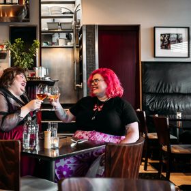 Two people enjoying a meal and clinking their wine glasses in Capitol Restaurant. Waitstaff make drinks behind the bar behind them. The dark wood tables add a moody vibe to the interior.