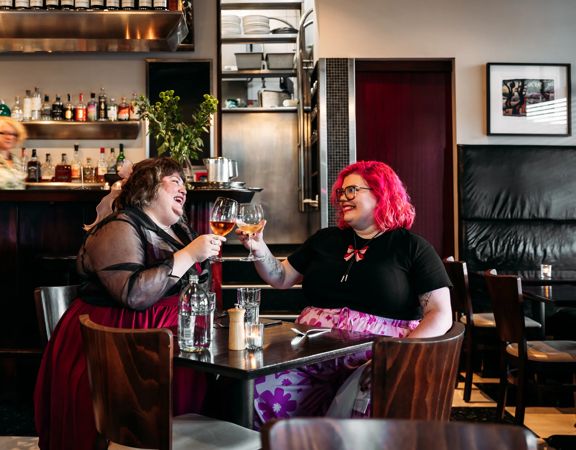 Two people enjoying a meal and clinking their wine glasses in Capitol Restaurant. Waitstaff make drinks behind the bar behind them. The dark wood tables add a moody vibe to the interior.