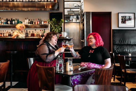 Two people enjoying a meal and clinking their wine glasses in Capitol Restaurant. Waitstaff make drinks behind the bar behind them. The dark wood tables add a moody vibe to the interior.