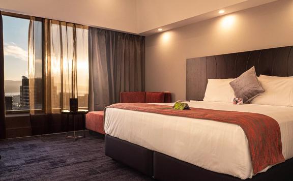 A room at the James Cook Hotel Grand Chancellor with a king-sized bed, beige walls, grey carpet, a red lounge chair and a large window with a view of Wellington. 